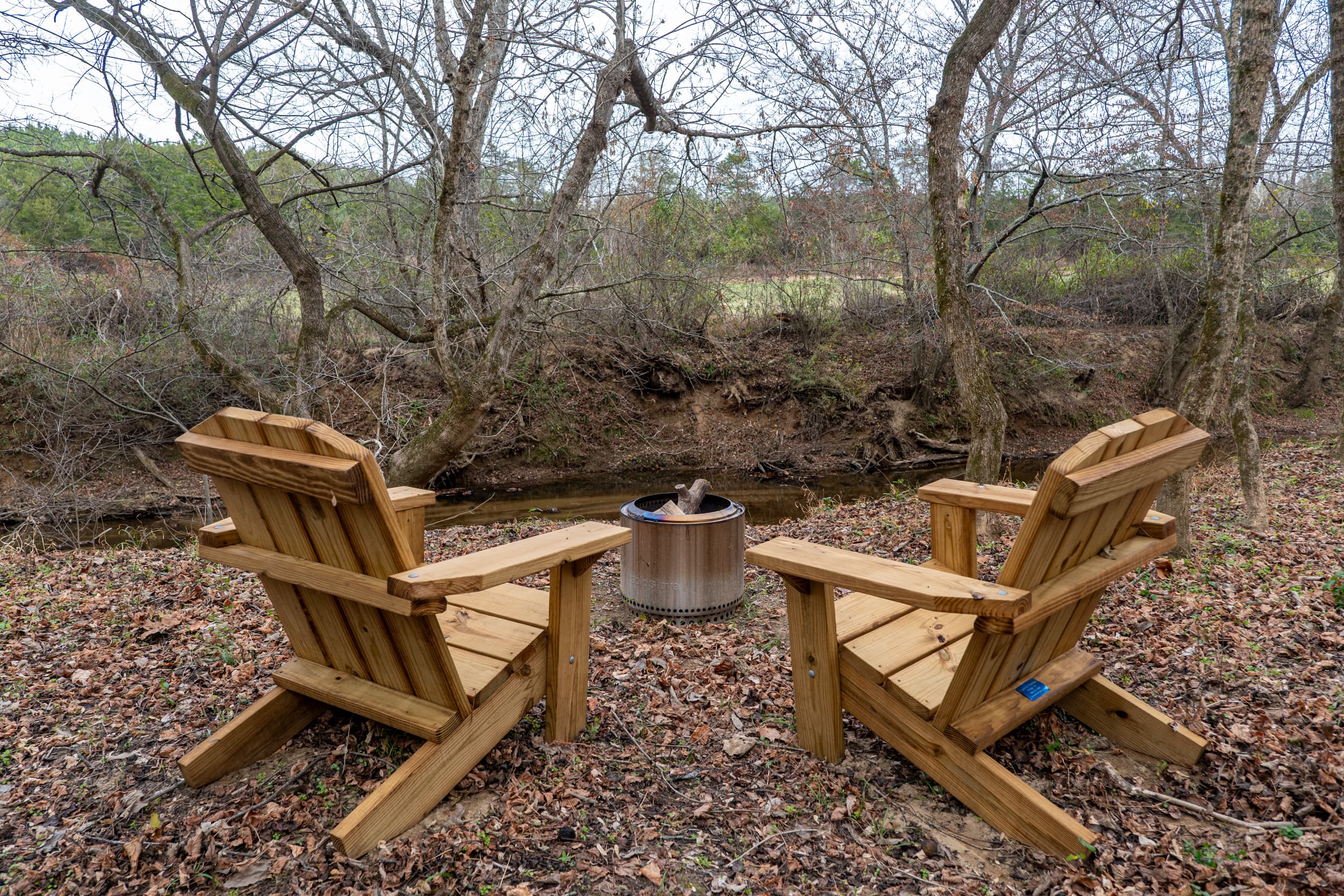 Two Adirondack chairs by the creek with firepit
